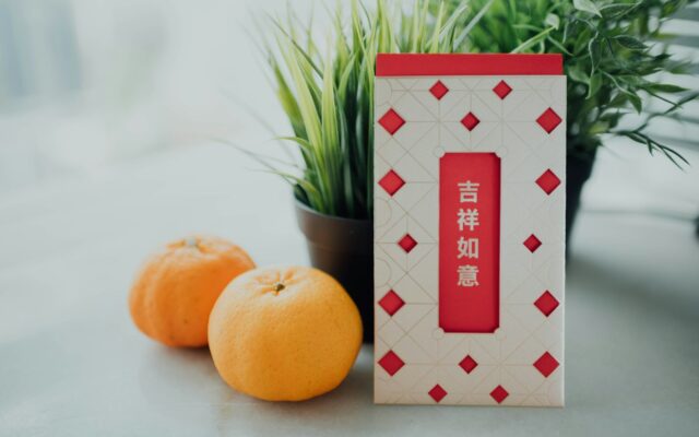 Red packet printed with 吉祥如意 beside two mandarin oranges
